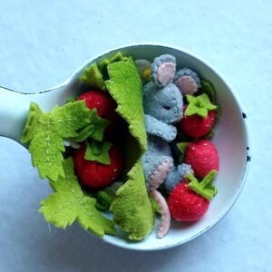 Op de afbeelding: Een grillige viltcreatie met een grijze muis genesteld tussen rode vilt aardbeien en groene vilt bladeren, allemaal in een witte, ondiepe kom. Het handgemaakte ontwerp roept een speelse, miniatuur voedselscène op.
