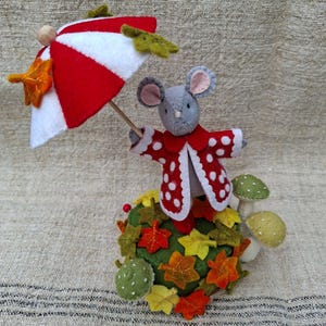 Könnte beinhalten: Eine Filzmausfigur in einem roten Mantel mit weißen Punkten, die einen rot-weiß gestreiften Regenschirm hält. Die Maus steht auf einem grünen Sockel, der mit Filz-Herbstblättern und drei kleinen grünen Pilzornamenten verziert ist.