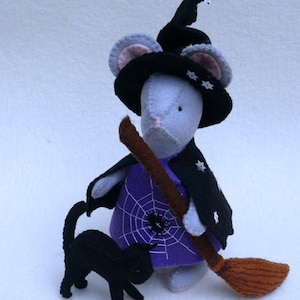 May include: A handmade Halloween-themed felt mouse dressed as a witch, wearing a black hat and cape with star accents. The mouse holds a brown broom and is accompanied by a black cat. The mouse wears a purple dress with a white spiderweb design.