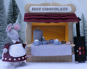 May include: A felt hot chocolate stall with the text "HOT CHOCOLATE" on a sign. A grey mouse in a red dress and white apron stands beside the stall. The stall has blue mugs, donuts, and a black stove.