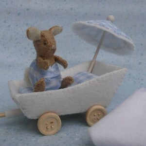Op de afbeelding: Een miniatuurviltmuis in een blauw-wit geruit outfit zit in een witte viltwagen met houten wielen. Een bijpassende parasol zorgt voor schaduw. De wagen wordt getrokken door een houten stok.