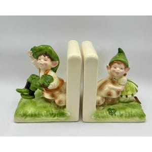 May include: A pair of ceramic bookends featuring whimsical elf figures. Each bookend has a cream-colored vertical support. The elves are depicted in green and brown attire, sitting on a green grassy base. One elf holds a green leafy item.