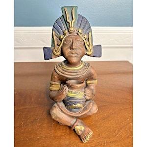 May include: A seated, hand-painted ceramic figurine of a person with a headdress, holding a small jar. The figurine is primarily brown with accents of blue, green, and gold. The jar has a blue and yellow design.