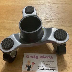 May include: A silver chair caster with four black wheels and three black rubber pads. The caster is designed to turn a chair into a rolling chair. The text on the card reads "Crafty Wheels Turn your chair into a rolling chair! Available in my Etsy shop: SassySissyofGeorgia"