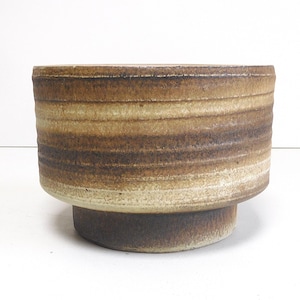Pode incluir: Um vaso cilíndrico de cerâmica com um exterior texturizado e canelado, apresentando faixas horizontais de esmalte em tons de castanho, bege e creme. O recipiente tem um corpo superior largo que se estreita para uma base circular mais estreita.