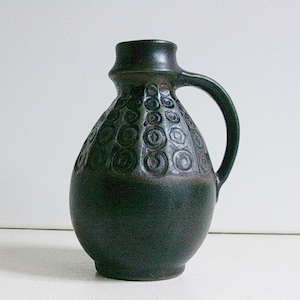 May include: A dark green ceramic vase with a circular pattern and a handle. The vase has a rounded body and a narrow neck.