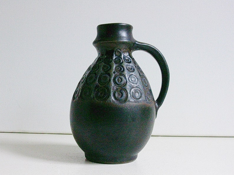 May include: A dark green ceramic vase with a circular pattern and a handle. The vase has a rounded body and a narrow neck.