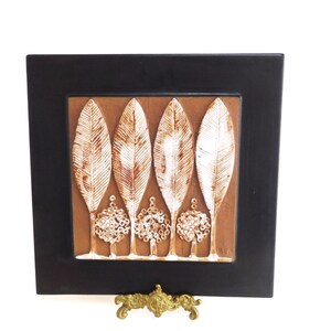 May include: A framed ceramic tile with a brown background and five white stylized trees. The trees are arranged in a row and have a leaf-like design. The tile is framed in black and is on a gold stand.