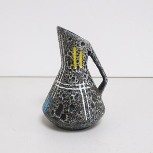 Peut inclure: Un petit vase en céramique gris avec une surface texturée. Le vase a une poignée et est décoré de lignes blanches et d'accents jaunes et bleus.