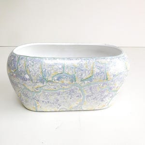 May include: An oval-shaped ceramic planter featuring a marbled exterior pattern in shades of light blue, lavender, and pale yellow with a crackled texture. The interior of the vessel is smooth and finished in a solid, clean white glaze.