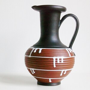 May include: A ceramic pitcher featuring a matte black neck and handle, contrasted with a ribbed, terracotta-colored body. The lower section is decorated with horizontal white lines and vertical white drip patterns that extend from the lines.