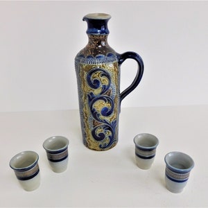 May include: A set of four small, white ceramic shot glasses with blue and gold trim, and a larger, blue and brown ceramic pitcher with a floral design.
