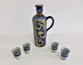 May include: A set of four small, white ceramic shot glasses with blue and gold trim, and a larger, blue and brown ceramic pitcher with a floral design.