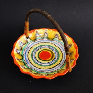 Puede incluir: Un plato de servir de cerámica colorido con un asa tejida. El plato tiene un borde festoneado y un diseño de círculos concéntricos en tonos de amarillo, verde, azul, rojo y blanco.