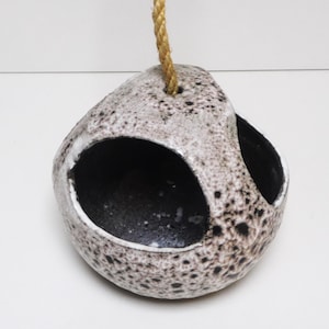 May include: A speckled ceramic hanging planter with two large openings and a natural rope for suspension. The planter has a neutral color palette with black and gray spots, creating a textured appearance. The interior is dark.