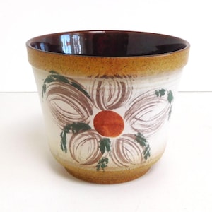 Op de afbeelding: Een bruine en witte keramische plantenpot met een gestileerd bloeiende ontwerp. De bloem is geschilderd in tinten van bruin, groen en oranje.