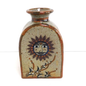 May include: A ceramic vase with a rustic brown and beige glaze. The vase features a sun with a face painted on the front. The sun is surrounded by brown and orange rays and has a blue centre. The vase has a small opening at the top.