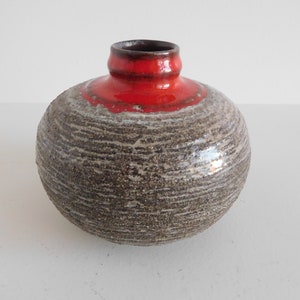 May include: A small, round ceramic vase with a textured, grey and white glaze. The top of the vase is a bright red colour.