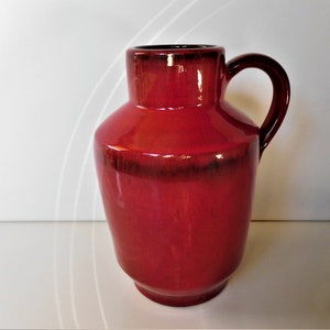 Può includere: Una brocca in ceramica rossa lucida con bordo e manico marrone scuro. La brocca ha un corpo arrotondato e un manico curvo. Anche l'interno della brocca è marrone scuro. È un oggetto decorativo.