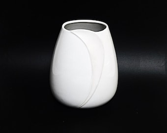 May include: A white ceramic vase with a unique, flowing design. The vase has a rounded, oval shape and a smooth, glossy finish.