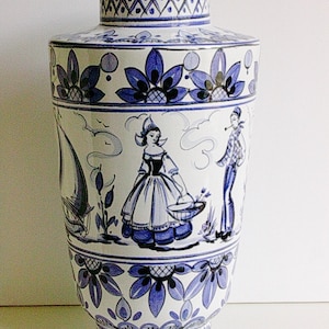 May include: A white ceramic vase with a blue and white floral design. The vase features a scene of two people in traditional Dutch clothing, one carrying a basket of flowers. The vase has a geometric pattern around the top and bottom.