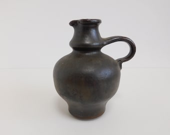 May include: A dark brown ceramic pitcher with a rounded body and a single handle. The pitcher has a textured surface and a slightly flared lip.