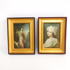 以下が含まれることがあります： 金色の縁取りのある額装された絵画が2つ。左の絵はピンクのドレスを着た女性と犬を描いています。右の絵は白い頭巾をかぶった女性を描いています。