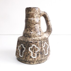 Puede incluir: Un jarrón de cerámica marrón y beige texturizado con asa. El jarrón presenta tres diseños de cruces blancas alrededor de la base. El jarrón tiene una estética rústica y vintage, adecuado para la decoración del hogar.