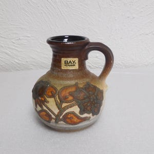 May include: A small, brown ceramic pitcher with a handle and a floral design. The pitcher has a dark brown rim and handle, with a beige and brown body. The floral design features orange and brown flowers and leaves. The label reads "BAY. KERAMIK."