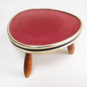 Puede incluir: Un pequeño reposapiés en forma de riñón con una superficie superior texturizada de color rojo oscuro. El borde está acabado con una banda metálica dorada acentuada por una fina raya negra. Se apoya en tres patas cónicas de madera clara.