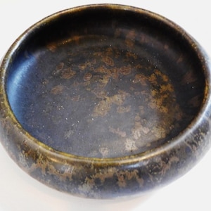 May include: A round ceramic bowl with a dark, mottled finish. The bowl's interior has a deep, blue-black hue with golden-brown speckles, and the rim has a contrasting bronze-green colour. Suitable for serving or display.