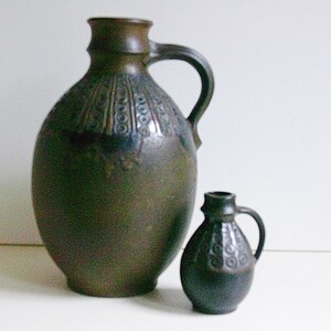 May include: Two brown ceramic jugs with a decorative pattern. The larger jug has a wide mouth and a handle. The smaller jug has a narrower mouth and a handle.