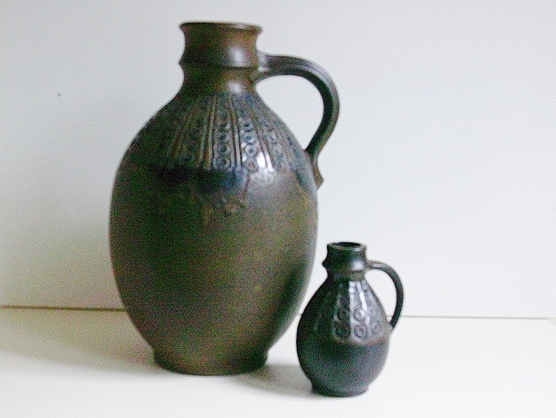 May include: Two brown ceramic jugs with a decorative pattern. The larger jug has a wide mouth and a handle. The smaller jug has a narrower mouth and a handle.