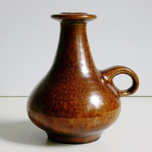 May include: A brown ceramic vase with a handle and a small opening. The vase has a bulbous body and a textured surface. The handle is a loop shape. The vase is approximately 18 cm tall.