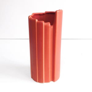 May include: A tall, terracotta-colored ceramic vase with a rectangular shape and vertical ridged detailing. The top edge has a scalloped design. The vase is set against a white background.