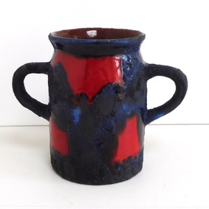 Puede incluir: Un jarrón de cerámica negro con dos asas y esmalte rojo y azul. El jarrón tiene una superficie texturizada.