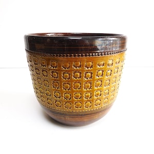 Op de afbeelding: Een vintage keramische plantenbak met een getextureerd, geometrisch patroon. De plantenbak heeft een donkerbruine rand en basis, met een warme, goudgele body. De body is versierd met een herhalend vierkant ontwerp.