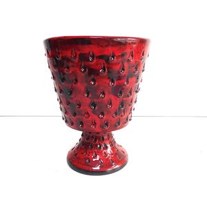 May include: A vibrant red ceramic chalice featuring a textured surface with raised, teardrop-shaped protrusions. The vessel has a wide, flared rim that tapers down to a narrow stem and a circular base, all finished in a glossy, mottled red glaze.