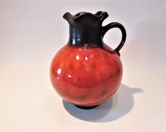 May include: A ceramic pitcher with a round body and a dark brown handle. The pitcher's body is a glossy red-orange color, transitioning to dark brown at the top. The rim has a wavy design.