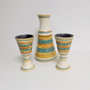 Op de afbeelding: Een keramische vaas en twee kelken. De set heeft een crèmekleurige basis met horizontale banden in bruin, teal en wit. De vaas heeft een hoge, slanke vorm, terwijl de kelken een uitlopende bovenkant en een voet hebben. Het ontwerp bevat bloemmotieven.