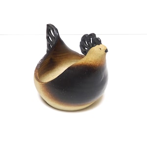 Puede incluir: Un cuenco de cerámica con forma de gallina, con cuerpo negro, cabeza beige y cresta y cola marrón oscuro. El cuenco tiene forma redondeada y está diseñado para guardar objetos pequeños. La gallina está sobre un fondo blanco.