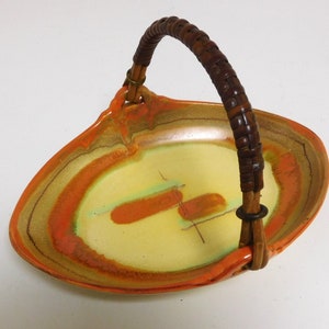 Puede incluir: Un plato de servir de cerámica con un asa tejida de color marrón. El plato tiene una base amarilla con vetas de color naranja y marrón.