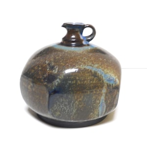 Op de afbeelding: Handgemaakte keramische kruik met een afgeronde body en een klein handvat. De kruik heeft een donkerbruin en blauw glazuur met een glanzende afwerking. De basis is donker en mat. Geschikt voor interieurdecoratie.
