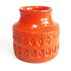 Op de afbeelding: Een feloranje keramische vaas met een getextureerd oppervlak en geometrische patronen. De vaas heeft een brede basis, een licht taps toelopend lichaam en een uitlopende rand. De decoratieve elementen omvatten ruit- en rechthoekvormen.