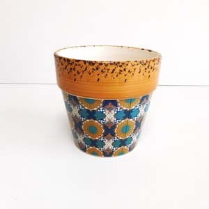 May include: A ceramic flower pot with a decorative floral pattern. The pot features a band of orange with a speckled texture at the top, and a blue, teal, and gold floral design on the body. The interior is white.