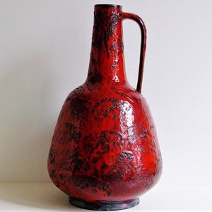 May include: A red ceramic vase with a textured surface and a handle. The vase has a wide base and a narrow neck.
