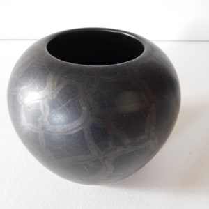 May include: A black ceramic vase with a round shape and a textured surface. The vase has a wide opening at the top.