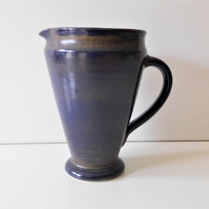 Op de afbeelding: Een donkerblauwe keramische kan met een gespikkeld glazuur. De kan heeft een brede basis en een afgeronde handgreep.