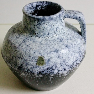 May include: A ceramic pitcher with a rounded body and a single handle. The surface features a mottled glaze pattern transitioning from a dark black base to a white and blue speckled top. A small, shield-shaped green sticker with text is visible.