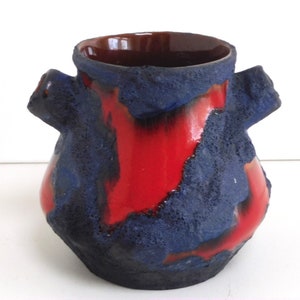 Puede incluir: Un jarrón de cerámica rojo y azul con una superficie texturizada y dos asas. El jarrón tiene la forma de un rectángulo redondeado.